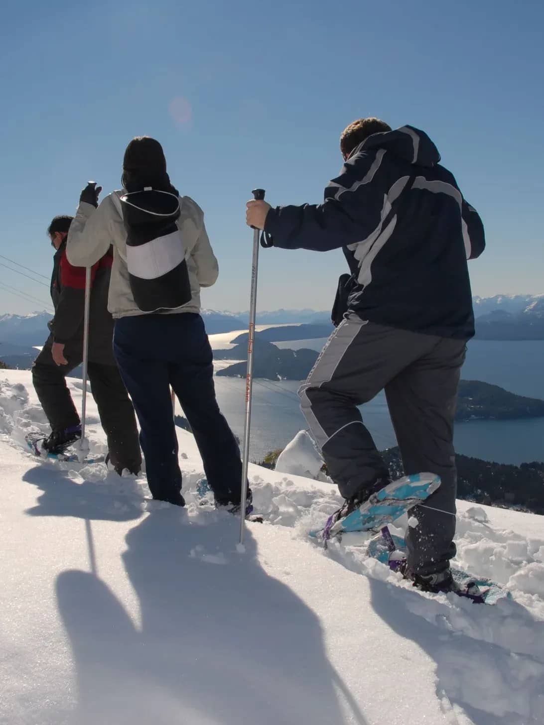 Three people walking with snowshoes accompanied by a guide on snow-covered ground with Lake Nahuel Huapi in the background