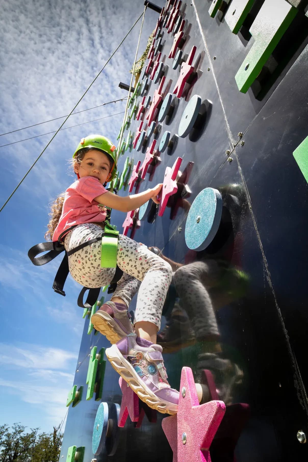 Una niña pequeña con casco verde y arnés de seguridad escala un muro de escalada lúdico al aire libre. El muro tiene presas de colores con formas de estrellas y círculos, y se ve un cielo azul con nubes ligeras de fondo desde un ángulo contrapicado.