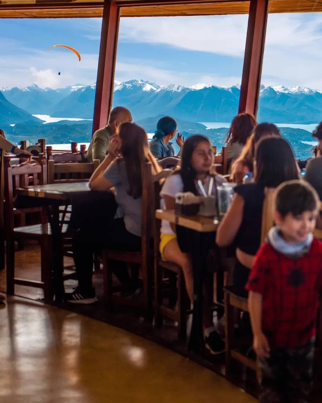 Gente almorzando en el interior de la Confiteria Giratoria con un fondo de montañas y el lago Nahuel Huapi que se ve a traves de la ventana